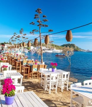 View of restaurant or cafe and bougainvillea flowers on beach in Gumusluk, Bodrum city of Turkey. Aegean seaside style colorful chairs, tables and flowers in Bodrum town near beautiful Aegean Sea.